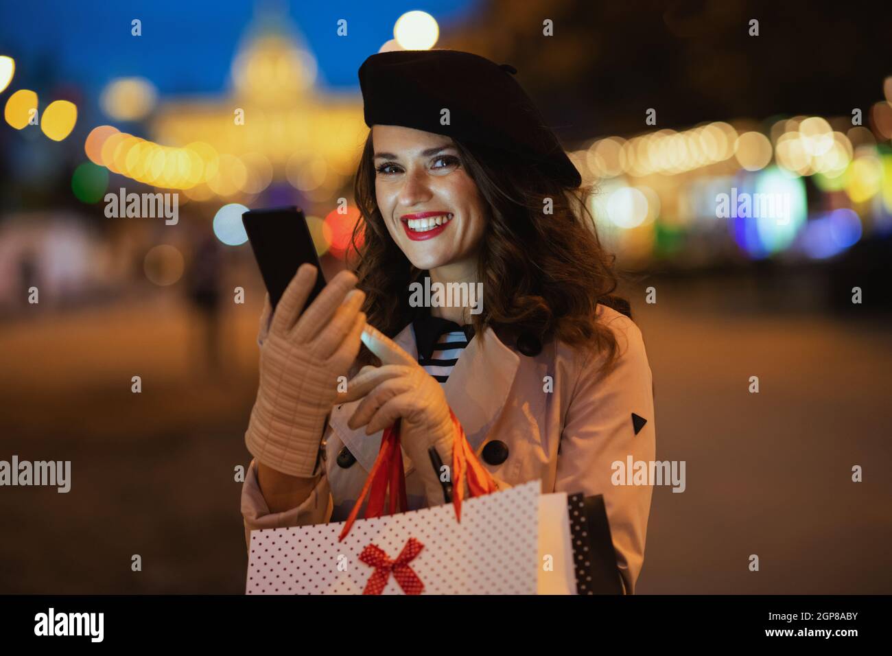Hello autumn. smiling elegant woman in beige trench coat with shopping bags sending text message using smartphone outside on the city street in autumn Stock Photo
