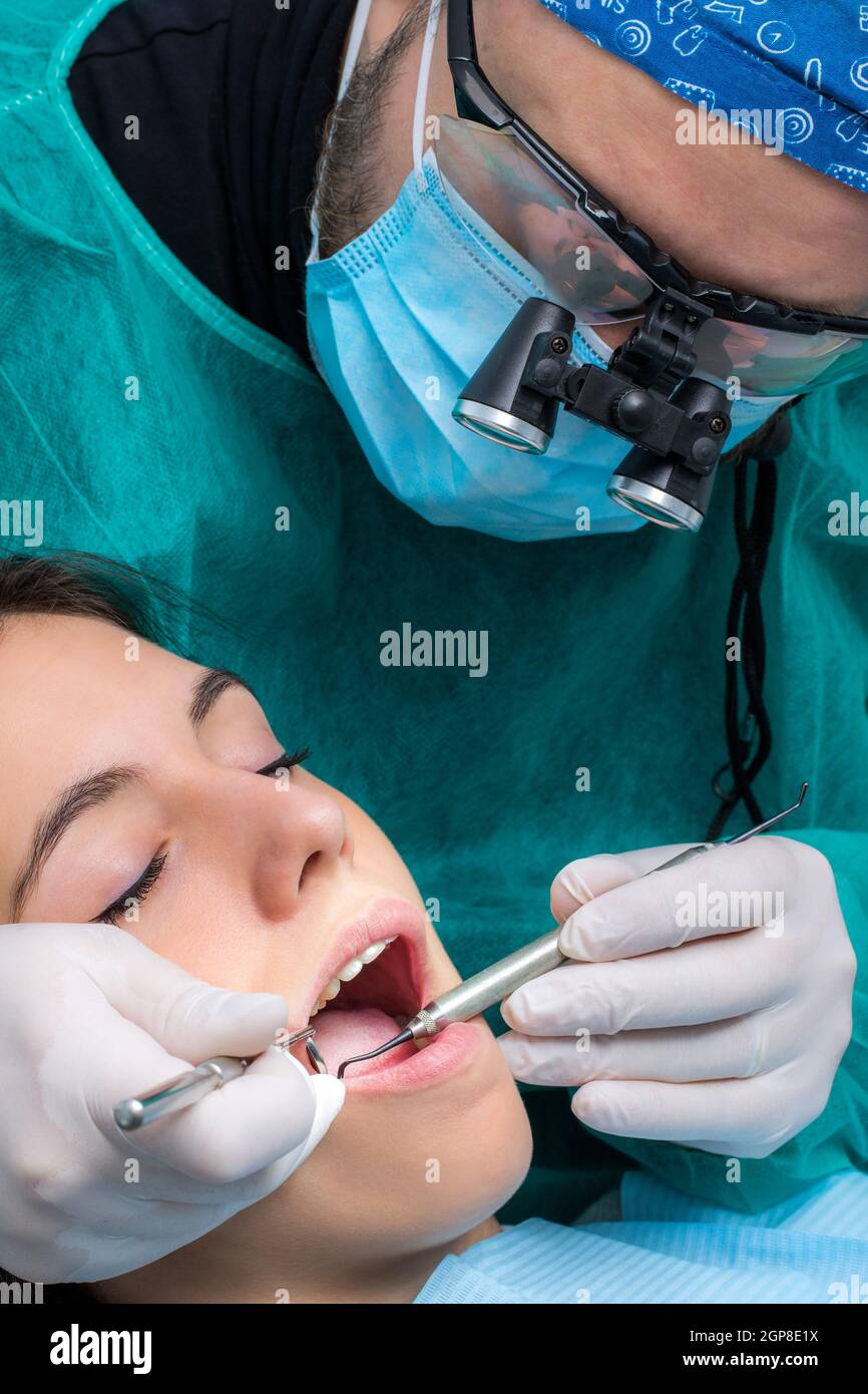 Close up of dentist wearing medical magnifying glasses working on female patient.Surgeon in green gown with mouth mirror and scaler. Stock Photo