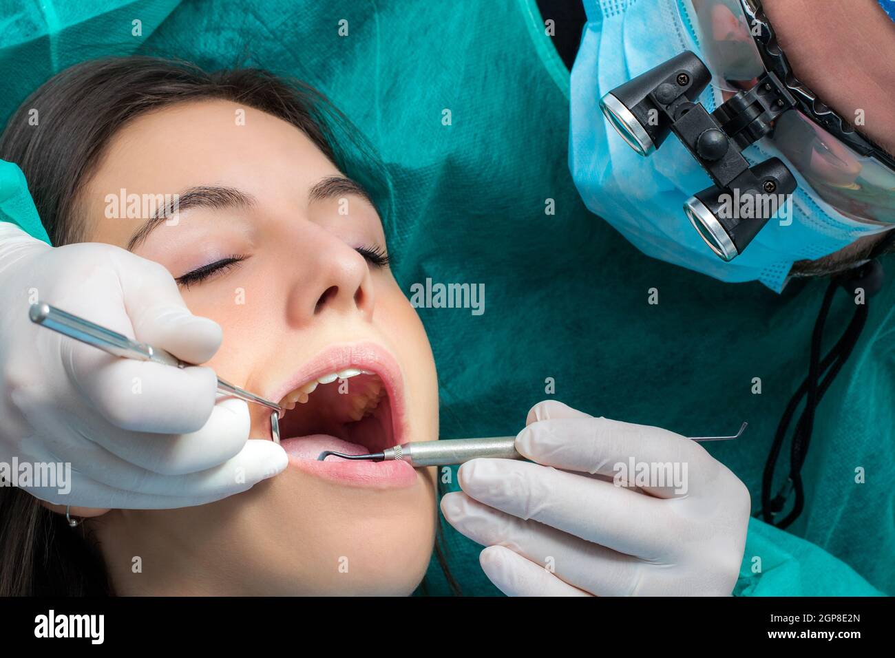 Close up of dentist wearing medical magnifying glasses working on female patient.Surgeon in green gown with mouth mirror and scaler. Stock Photo
