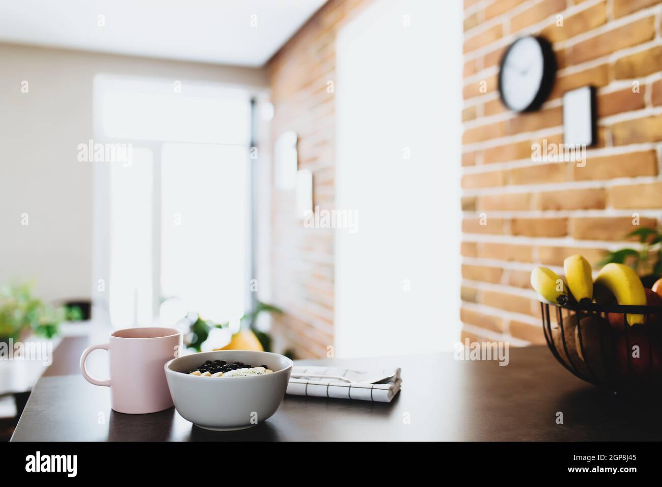 Pink coffee cup, bowl with chopped tropical fruits kiwi and banana, blueberries, spoon on bar counter in stylish loft kitchen. Blurred background. Hig Stock Photo