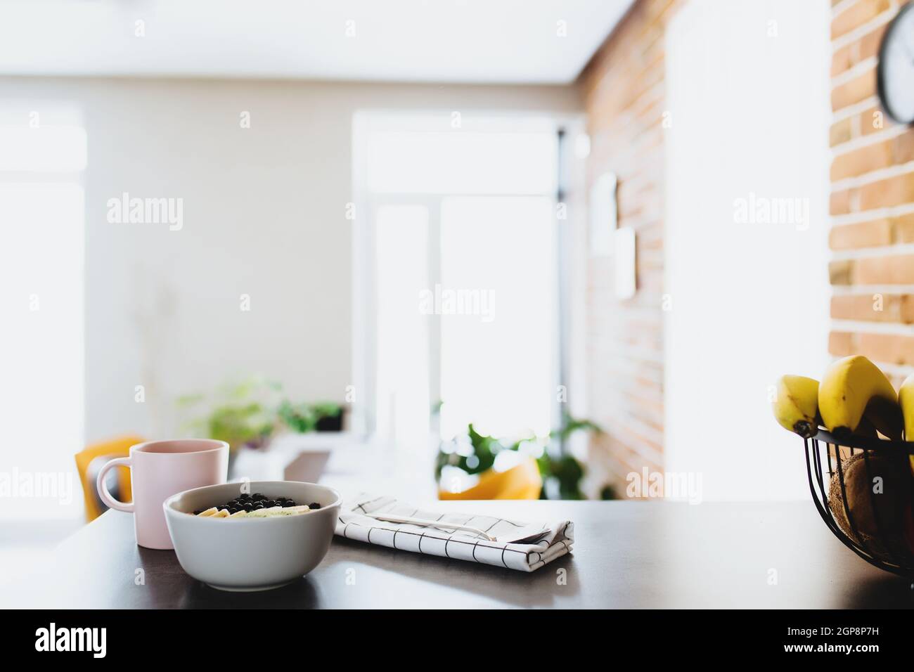 Pink coffee cup, bowl with chopped tropical fruits kiwi and banana, blueberries, spoon on bar counter in stylish loft kitchen. Blurred background. Hig Stock Photo