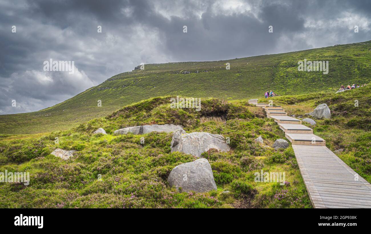 People hiking on wooden boardwalk, between boulders and heathers, leading to Cuilcagh Mountain. Storm, dramatic sky in background, Northern Ireland Stock Photo