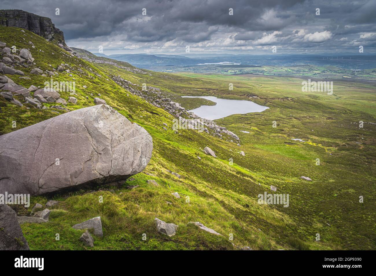 Large boulder and rubble on Cuilcagh Mountain mountainside, small lake in valley below and dramatic, stormy sky in background, Northern Ireland Stock Photo