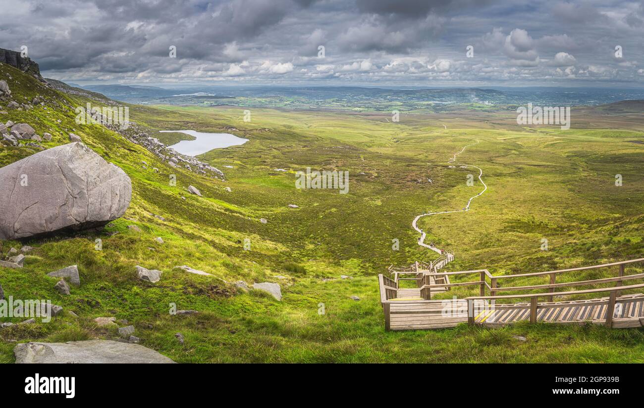 Large panorama with view on steep stairs of wooden boardwalk leading to Cuilcagh Mountain peak with lake and valley or plane below, Northern Ireland Stock Photo