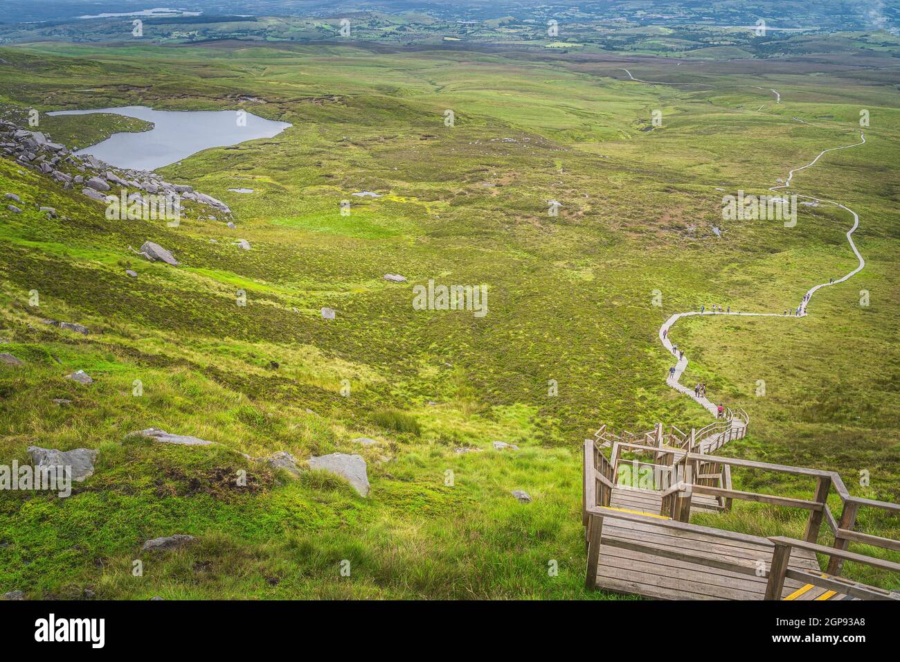 People climbing on steep steps and stairs of wooden boardwalk, to reach Cuilcagh Mountain peak with view on lake and valley below, Northern Ireland Stock Photo