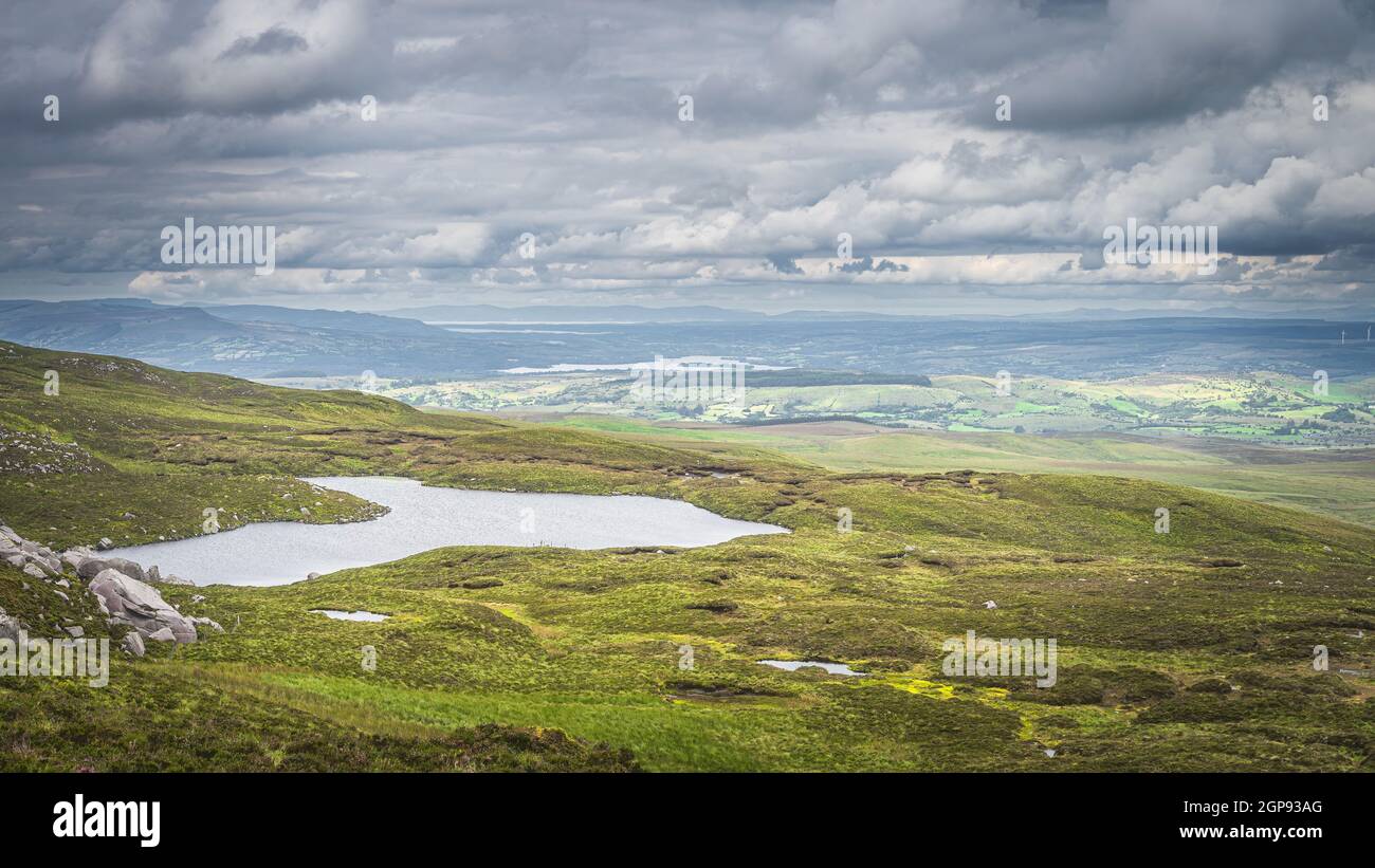 Green fields or rolling hills with small lake at footstep of Cuilcagh Mountain, dramatick stormy sky in background, Northern Ireland Stock Photo