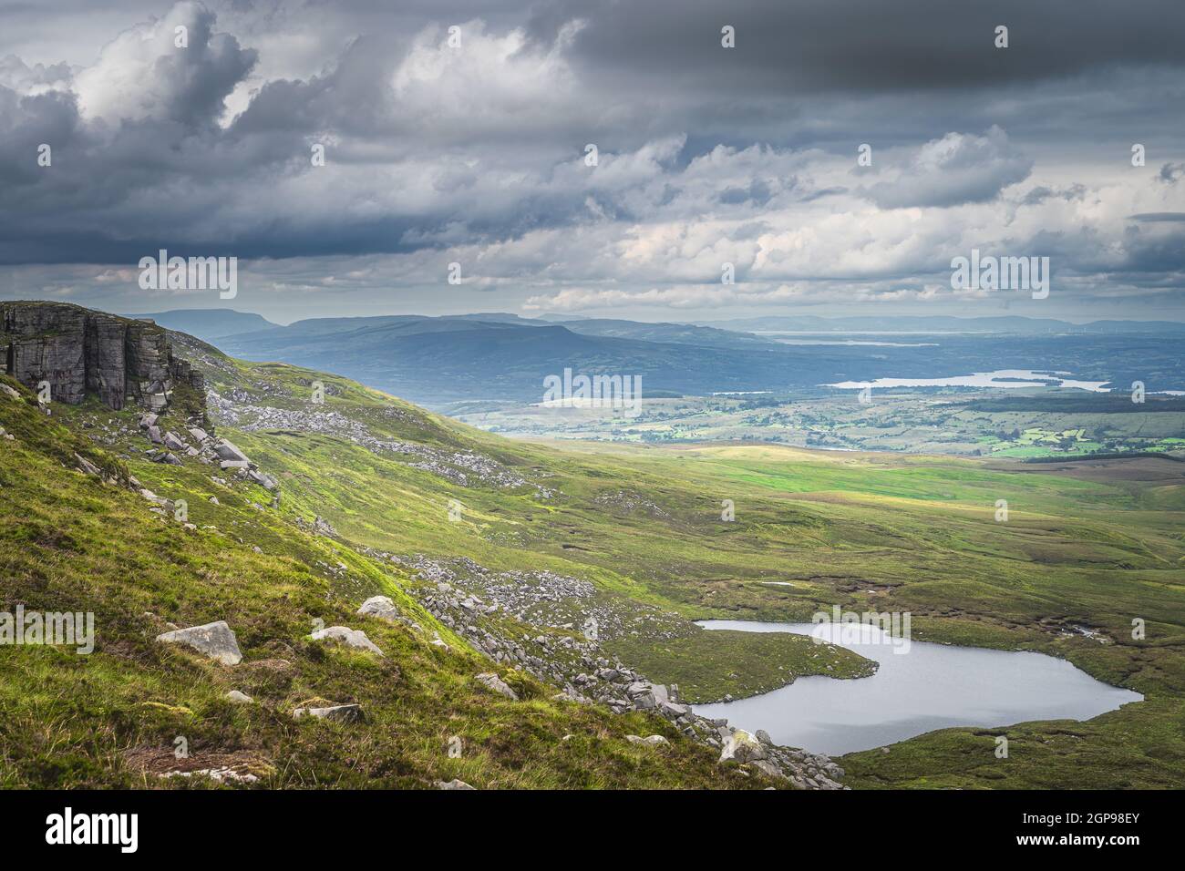 Green fields and large boulders illuminated by sunlight in Cuilcagh Mountain Park with small lakes in valley below, Northern Ireland Stock Photo