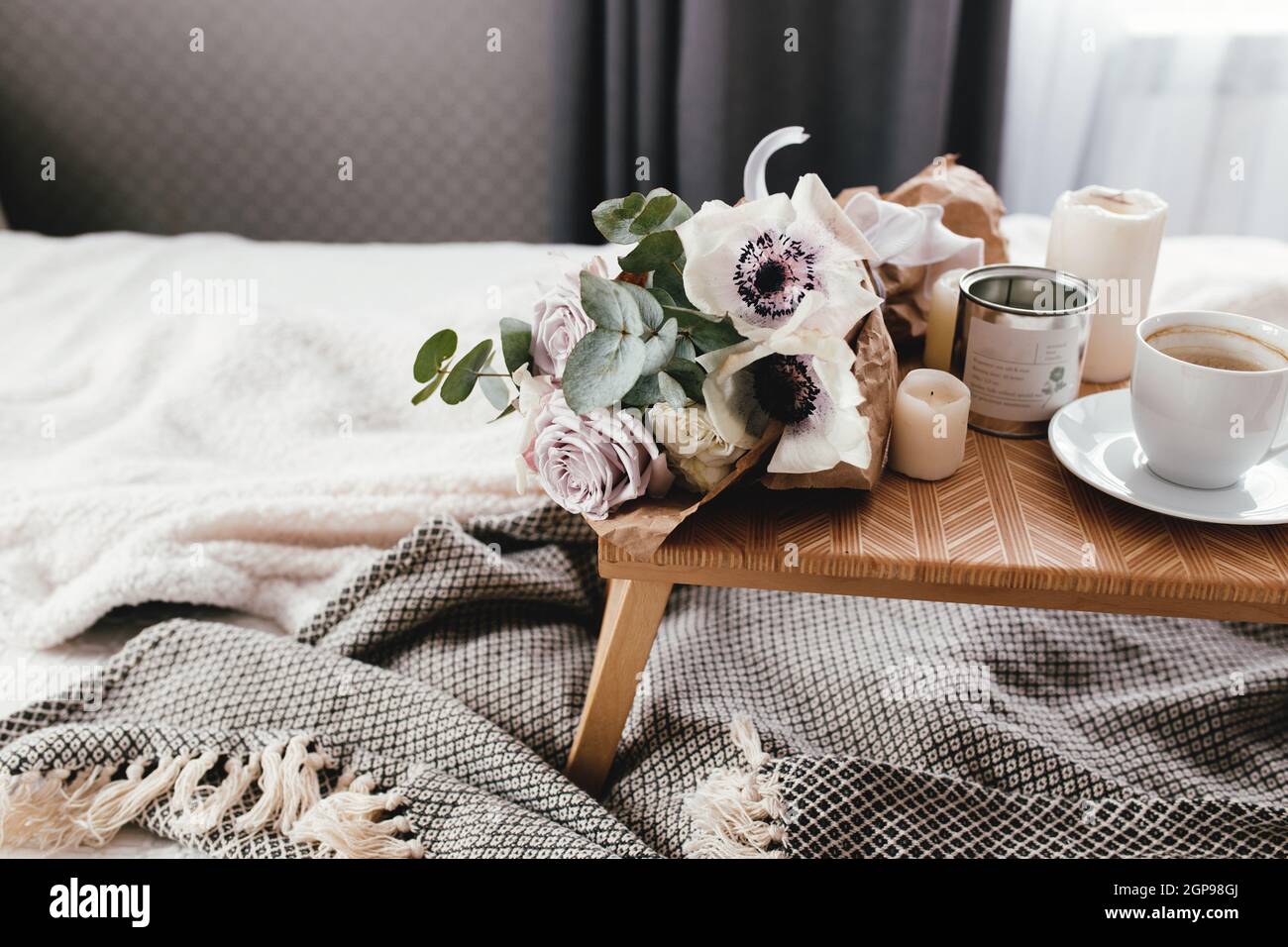 Romantic morning. Wooden coffee table with flowers on bed with plaid, coffee cup, flowers and candles. Lilac roses with eucalyptus and anemones. Inter Stock Photo