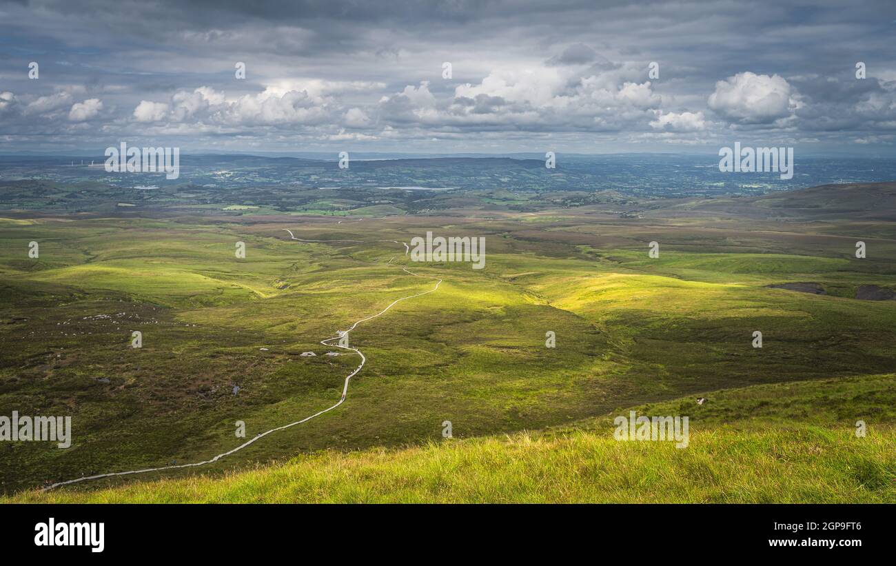 Winding wooden path of Cuilcagh Mountain Park boardwalk, illuminated by patches of sunlight in the valley below with dramatic sky, Northern Ireland Stock Photo