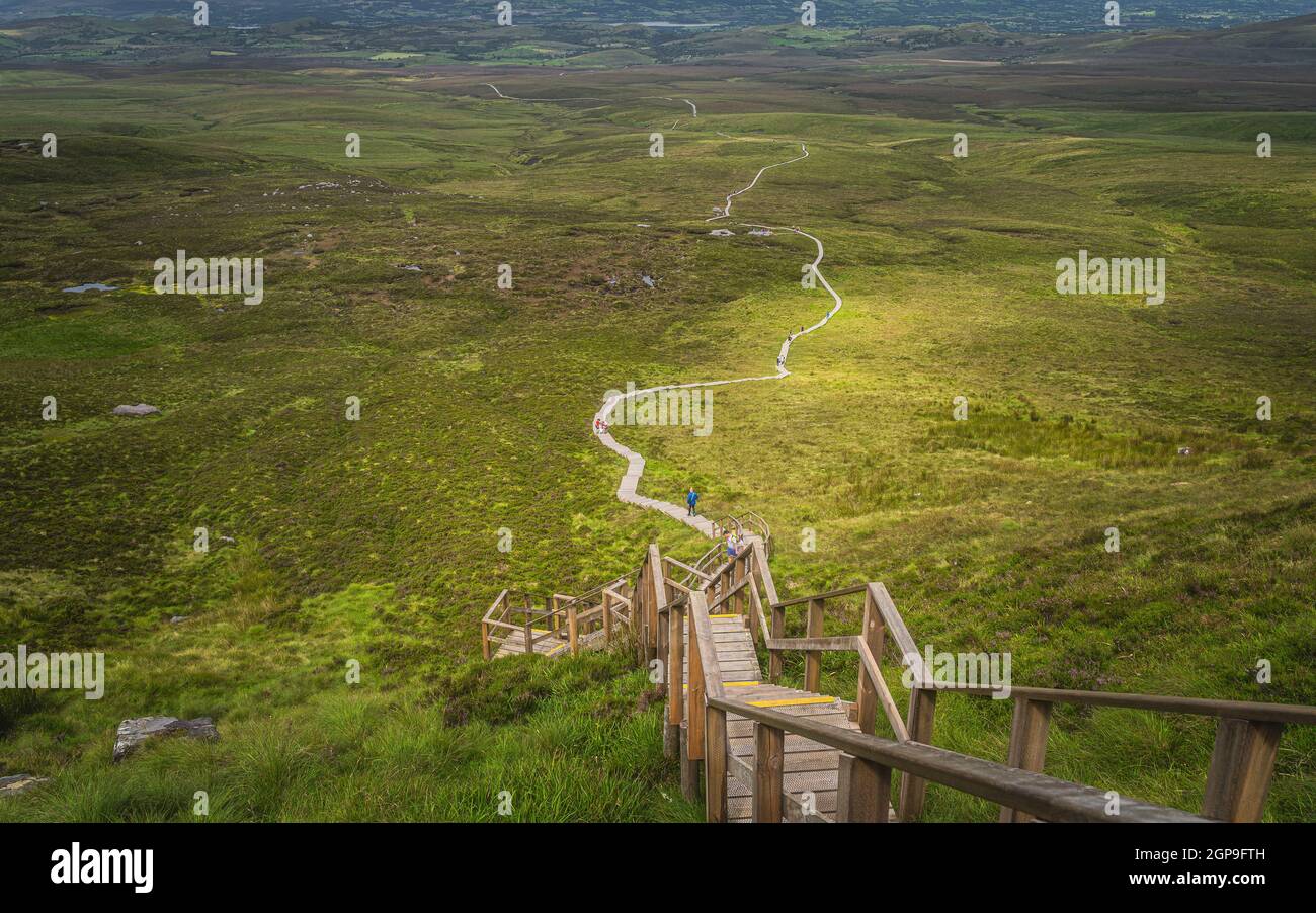 People climbing on steep stairs of wooden boardwalk, to reach Cuilcagh Mountain peak with view on green fields and valley below, Northern Ireland Stock Photo