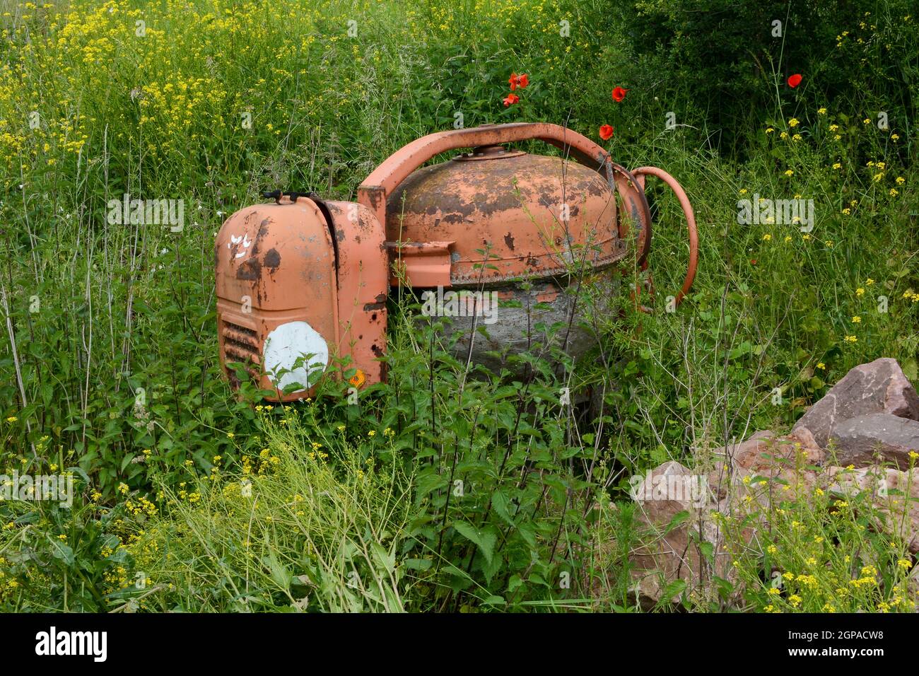 Old rusted cement mixer in the middle of a tall green meadow with big stones Stock Photo