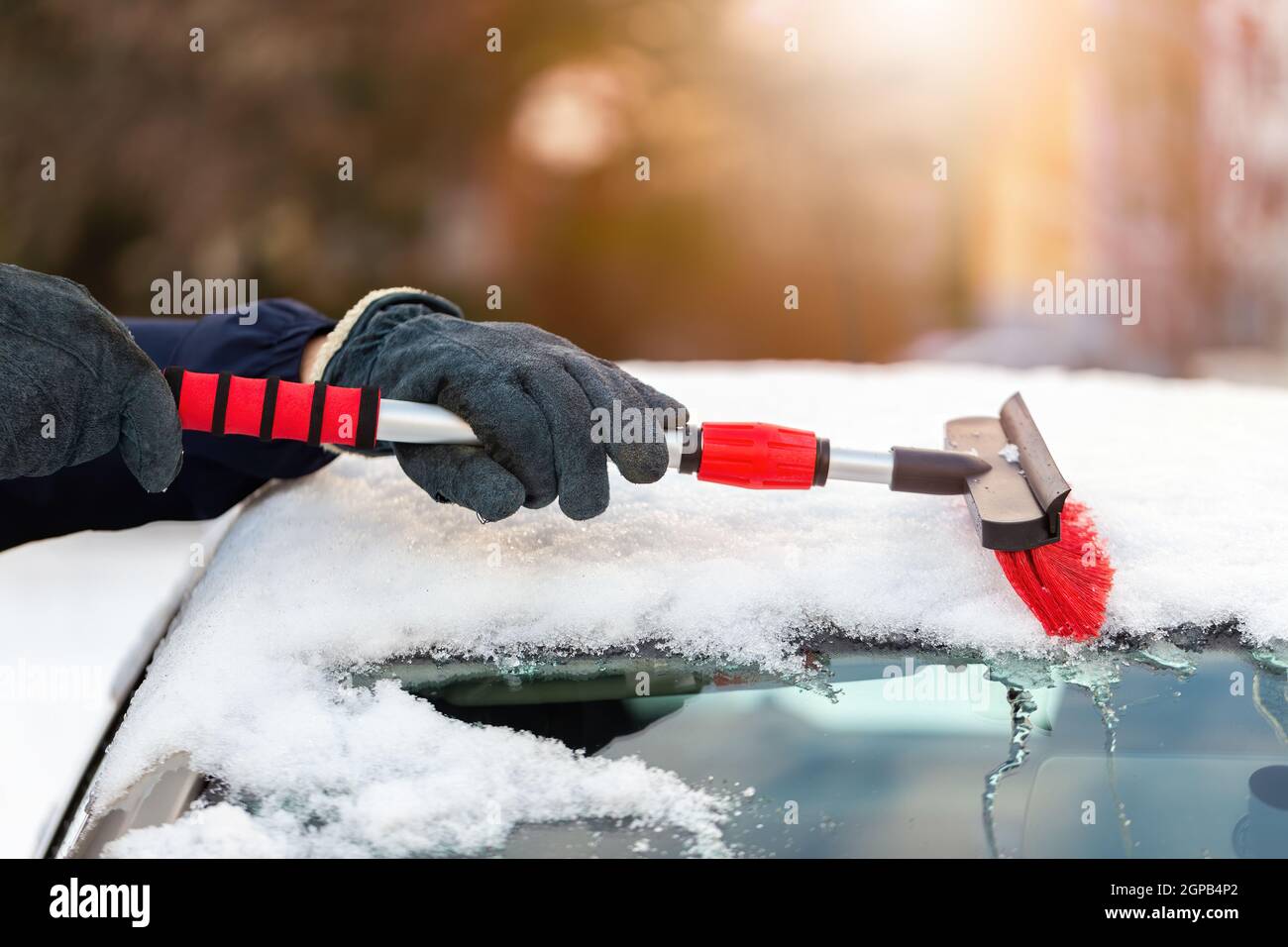 Person in gloves sweeping snow from the car in sunny day. Human cleaning snowy cover from automobile with broom. Scraping frost from roof of the vehic Stock Photo