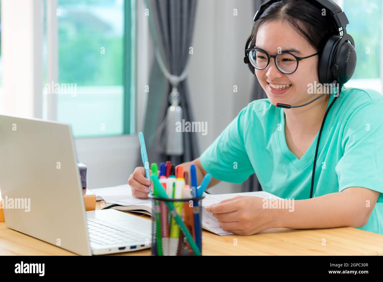 Asian young woman student with glasses headphones girl study happy writing note on a book looking video conference laptop computer university class on Stock Photo