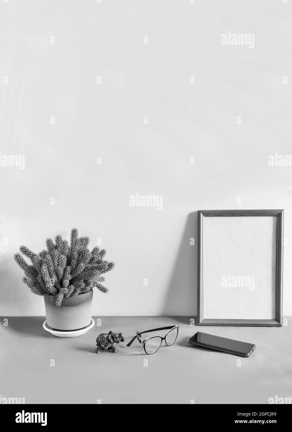 On a table against a white wall is a wooden photo frame and a potted cactus flower. Next to it are glasses and a smartphone. Front view, copy space Stock Photo