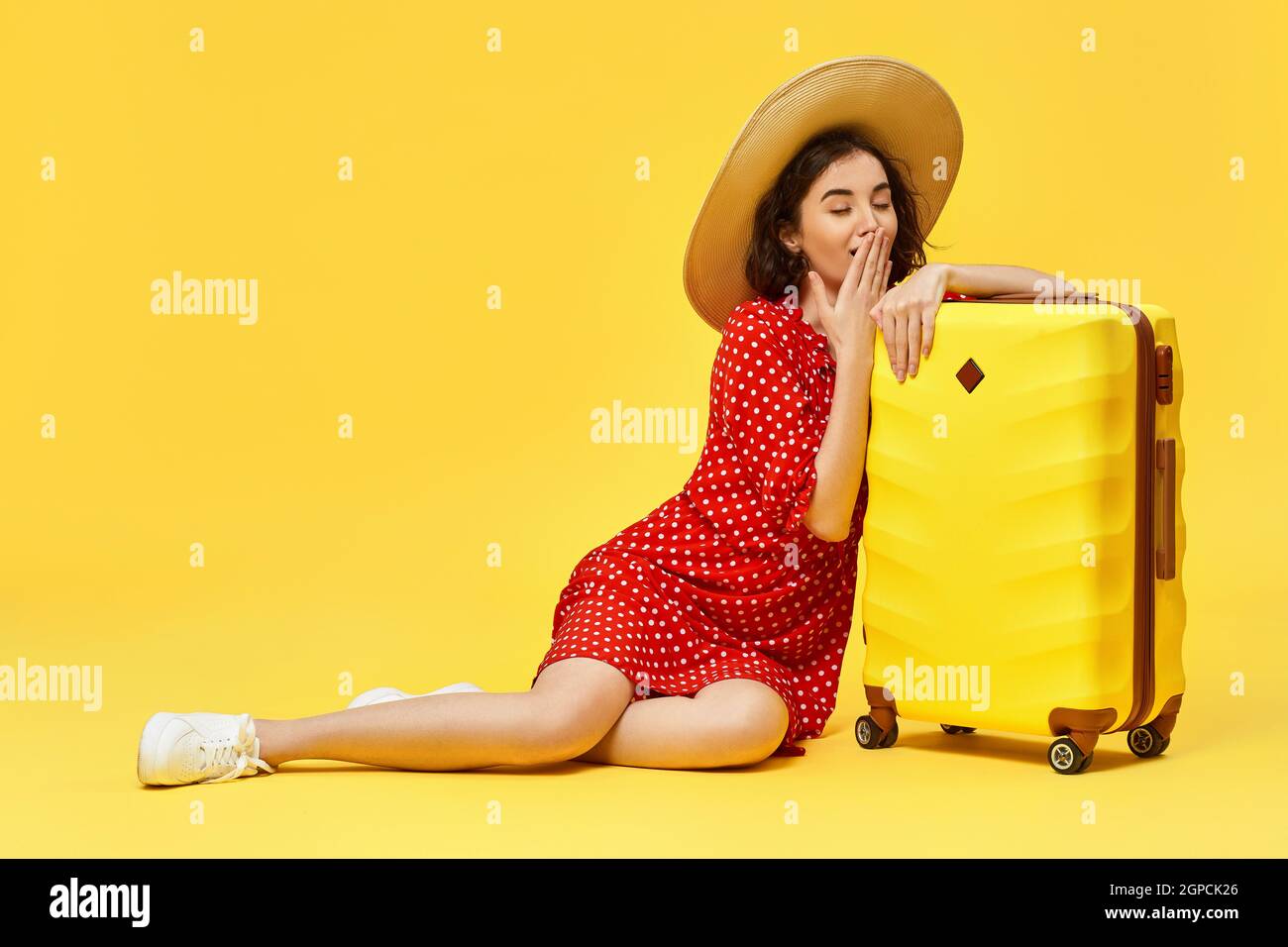 Happy woman in red dress with suitcase waiting for her flight on yellow background. flight delay and cancelled travel Stock Photo