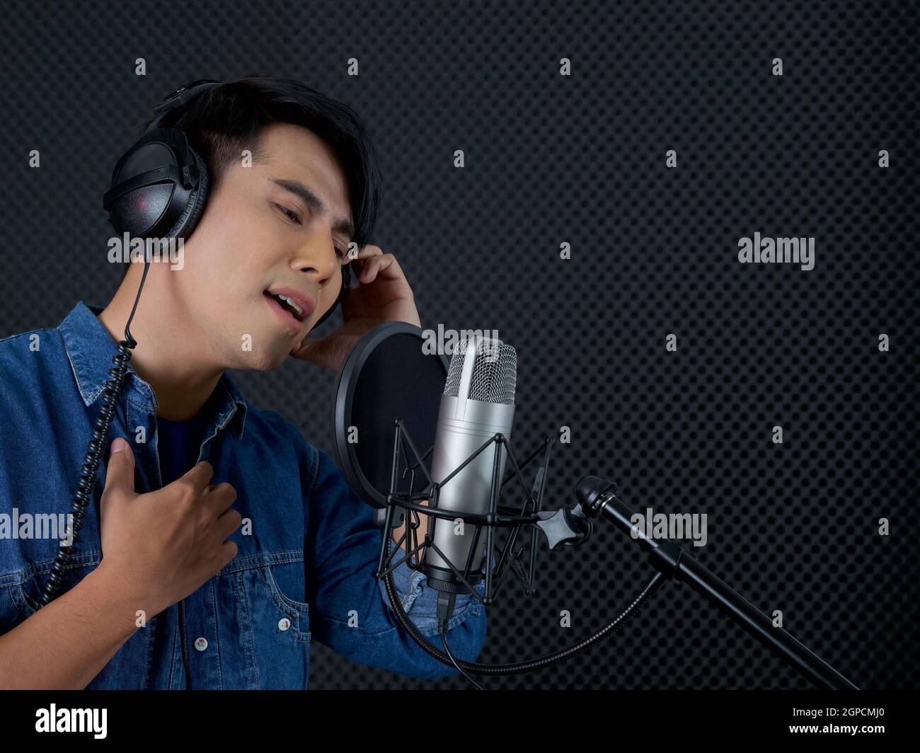 Young asian man with headphone  singing in front of black soundproofing wall. Musician producing music in professional recording studio. Stock Photo