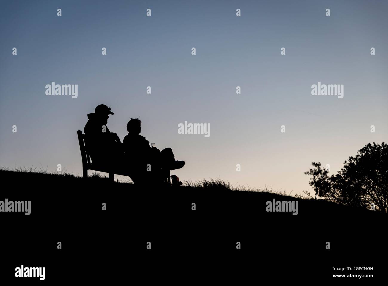 A couple sitting on a park bench, silhouetted against a dusk sky, picnicing and enjoying the sunset Stock Photo