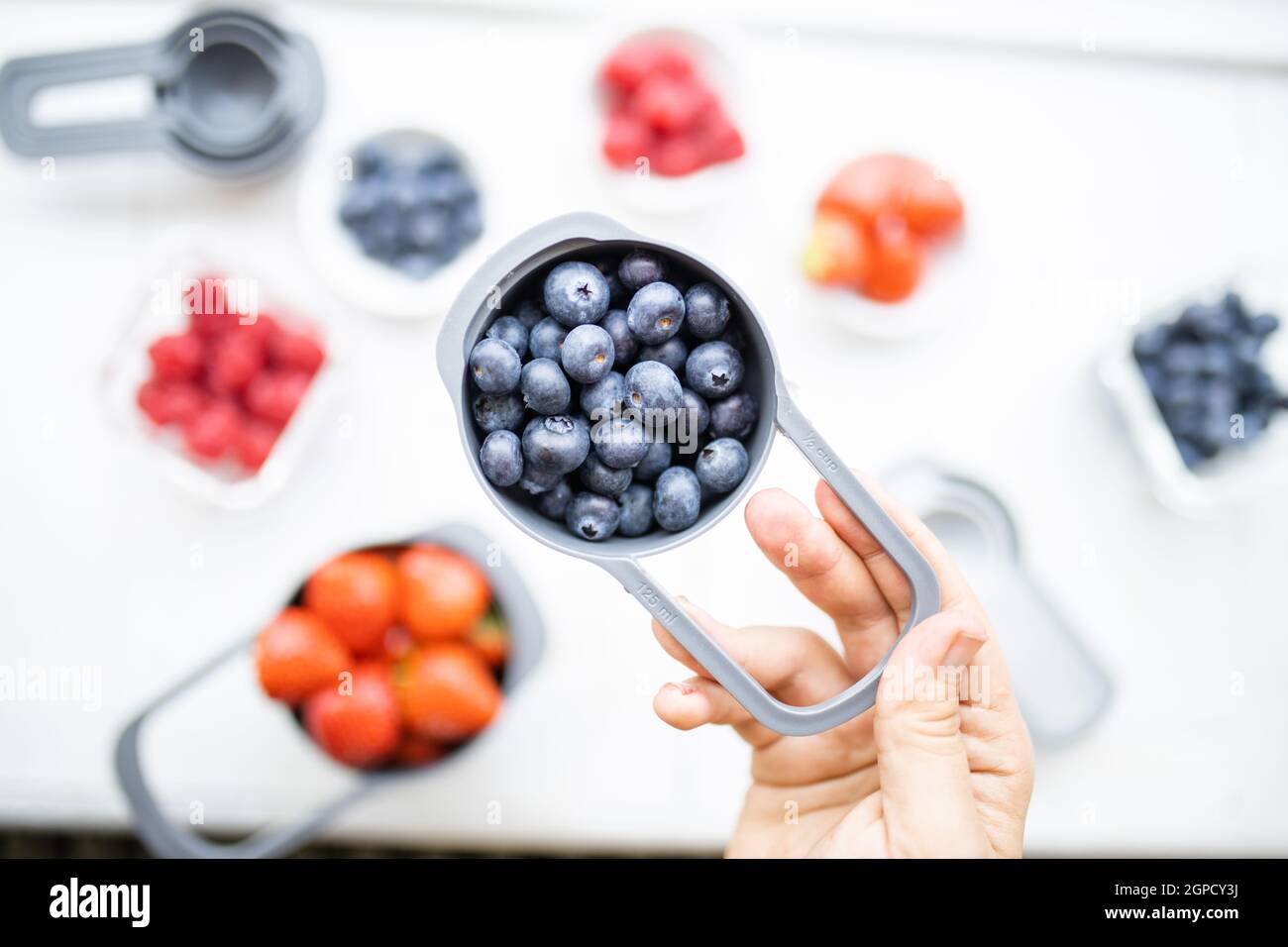 Female hand holding measuring cup of blueberries above more berries in containers. Blueberries, raspberries, and strawberries on table from above. Fru Stock Photo