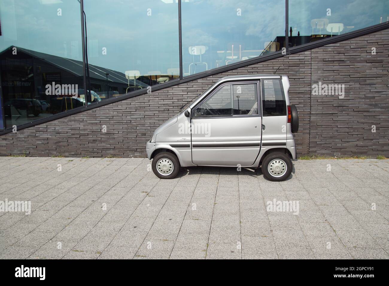 Gray microcar Canta in front of a gray building with many large windows in the Dutch city of Alkmaar. Netherlands, September Stock Photo