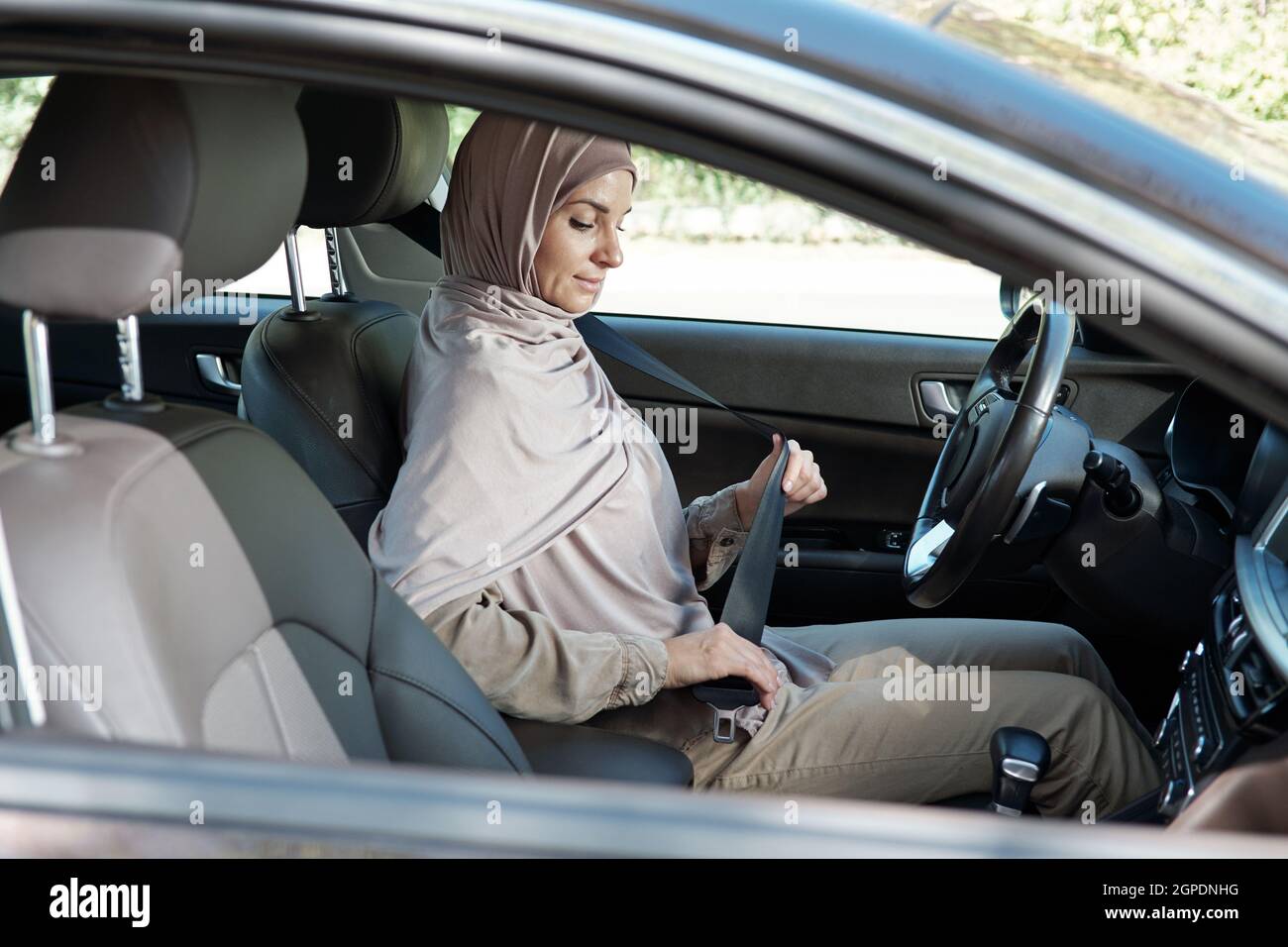 Horizontal shot of modern Muslim woman wearing hijab sitting in her car buckling seatbelt before starting driving Stock Photo