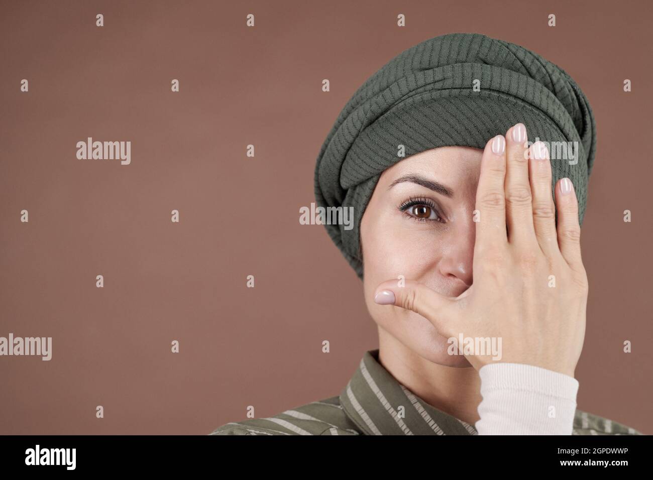 Horizontal close-up studio portrait of attractive Muslim woman wearing stylish shirt and turban covering half of face with hand looking at camera, brown background Stock Photo