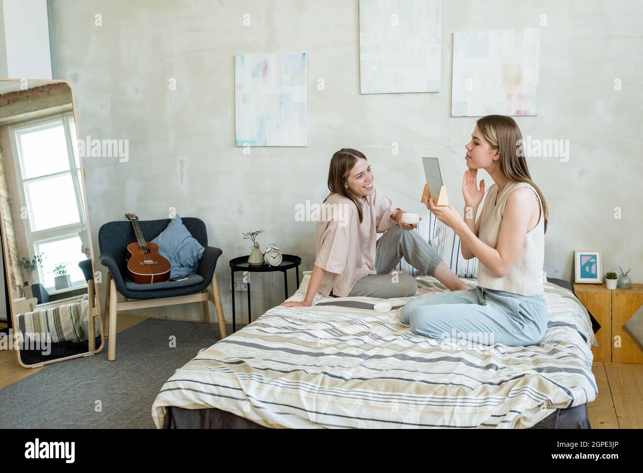 Happy girl with drink looking at her twin sister taking care of her skin while both relaxing on bed Stock Photo