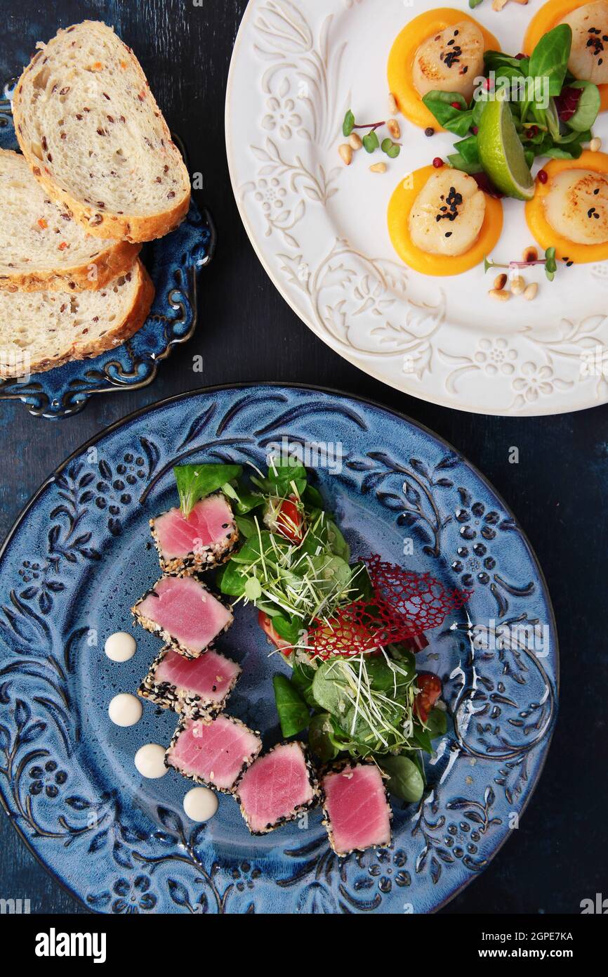 Slices of tuna in sesame seeds on a blue ceramic plate.Scallop with pumpkin puree with micro greens and pine nuts on a white ceramic plate.Vertical ph Stock Photo