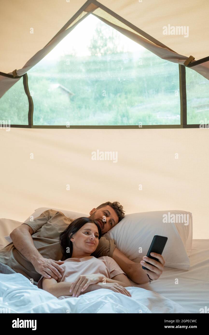 Young restful couple with smartphone making video call while relaxing in bed inside glamping house Stock Photo