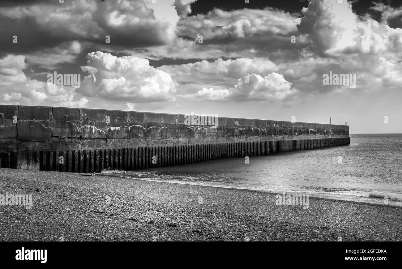 Black and white image of lone fisherman at Shoreham-by-sea at end of jetty wall with beach and sea. Stock Photo