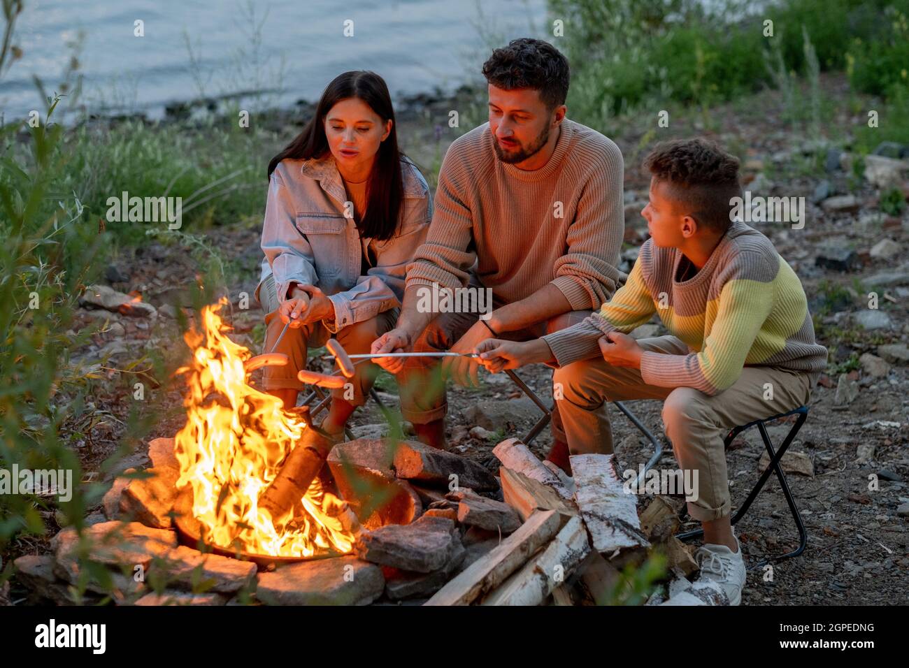 Three young backpackers frying sausages on campfire in the evening Stock Photo