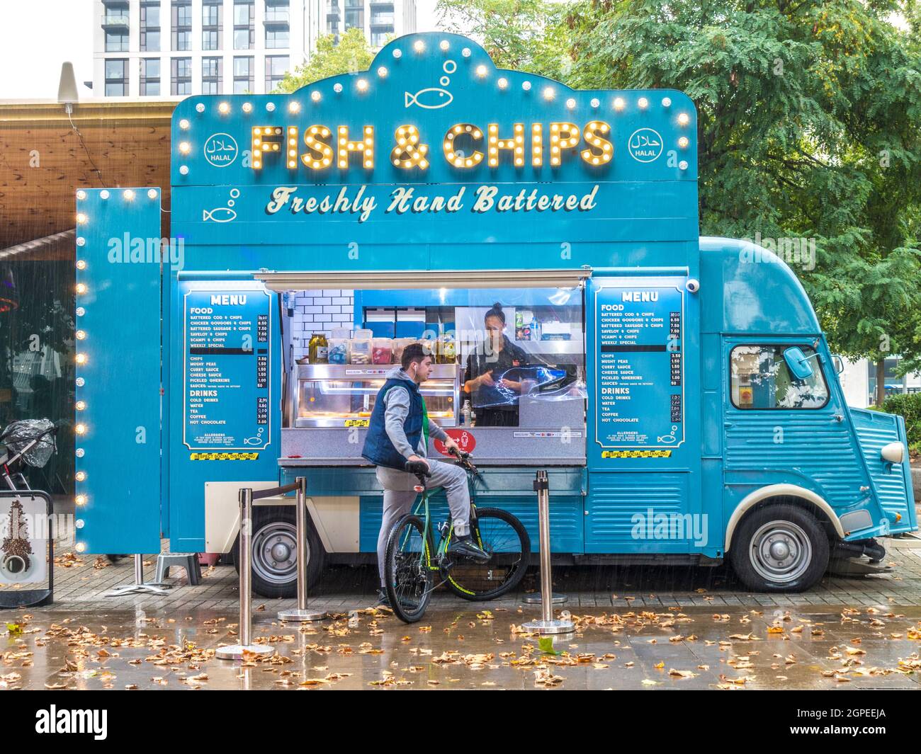 A man on a bicycle waiting in autumn rain for food to be served by a lady at a converted Fish & Chips van, parked at South Bank, London, England, UK. Stock Photo