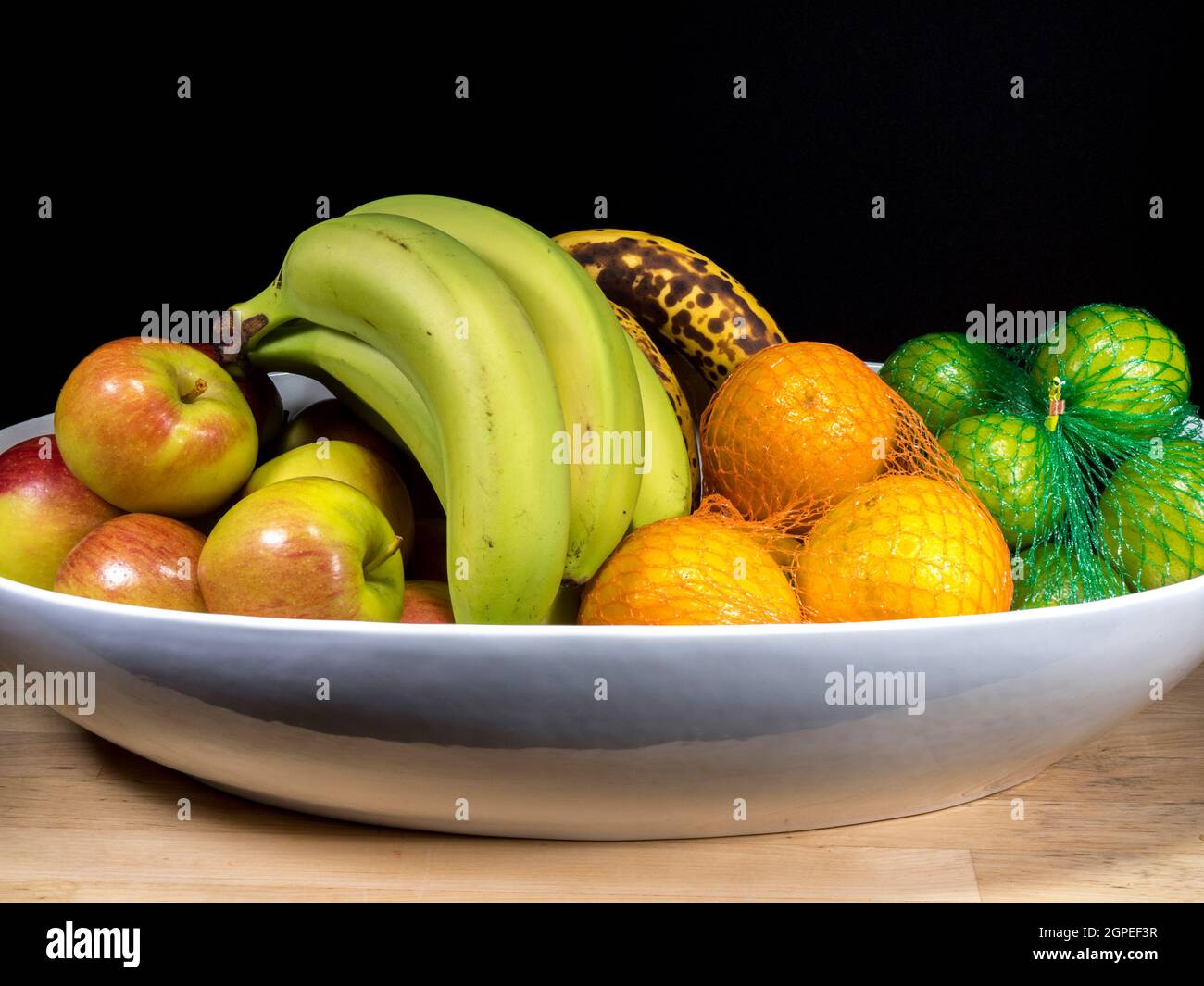 Closeup POV isolated shot of oranges and limes in mesh bags next to apples and bananas in a white display bowl. Stock Photo