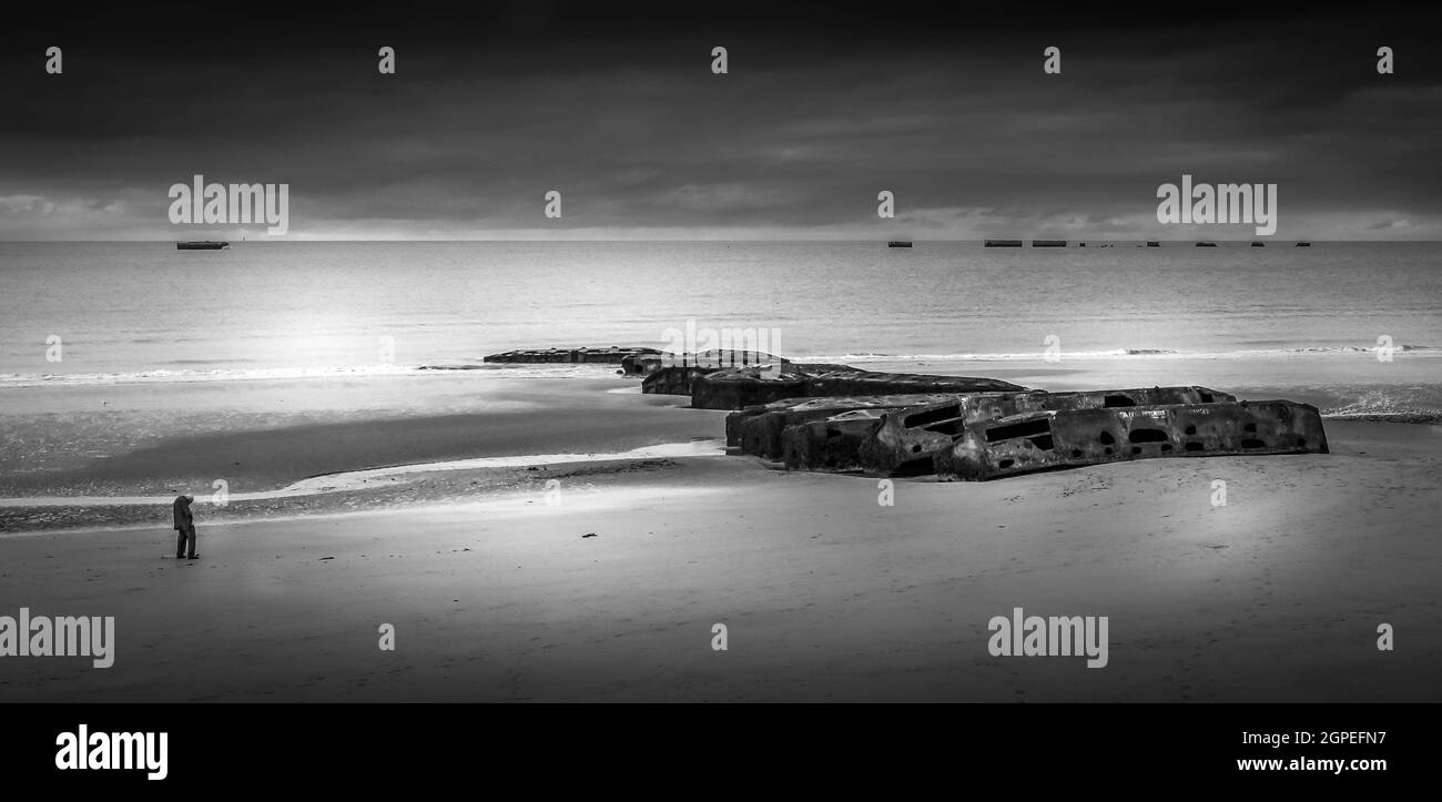 Black and white image of lone person remembering the 6th June 1944. with mulberry harbour remains on beach. Stock Photo