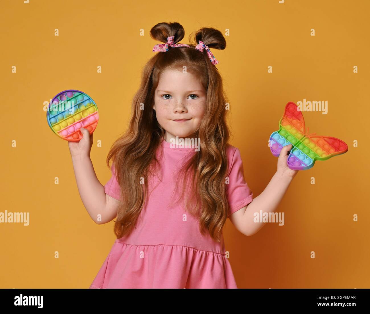 Smiling red-haired kid girl in pink clothes holds two new sensory rainbow color round and butterfly shape toy - pop it Stock Photo