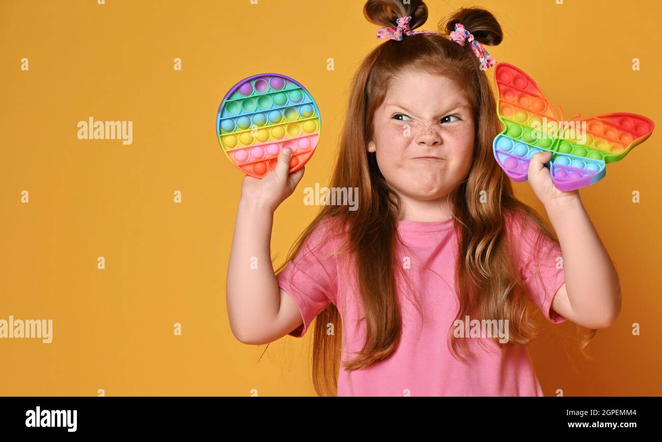Frolic grimacing kid girl in pink shirt holds two sensory rainbow color toys - pop it and looks aside at round one Stock Photo