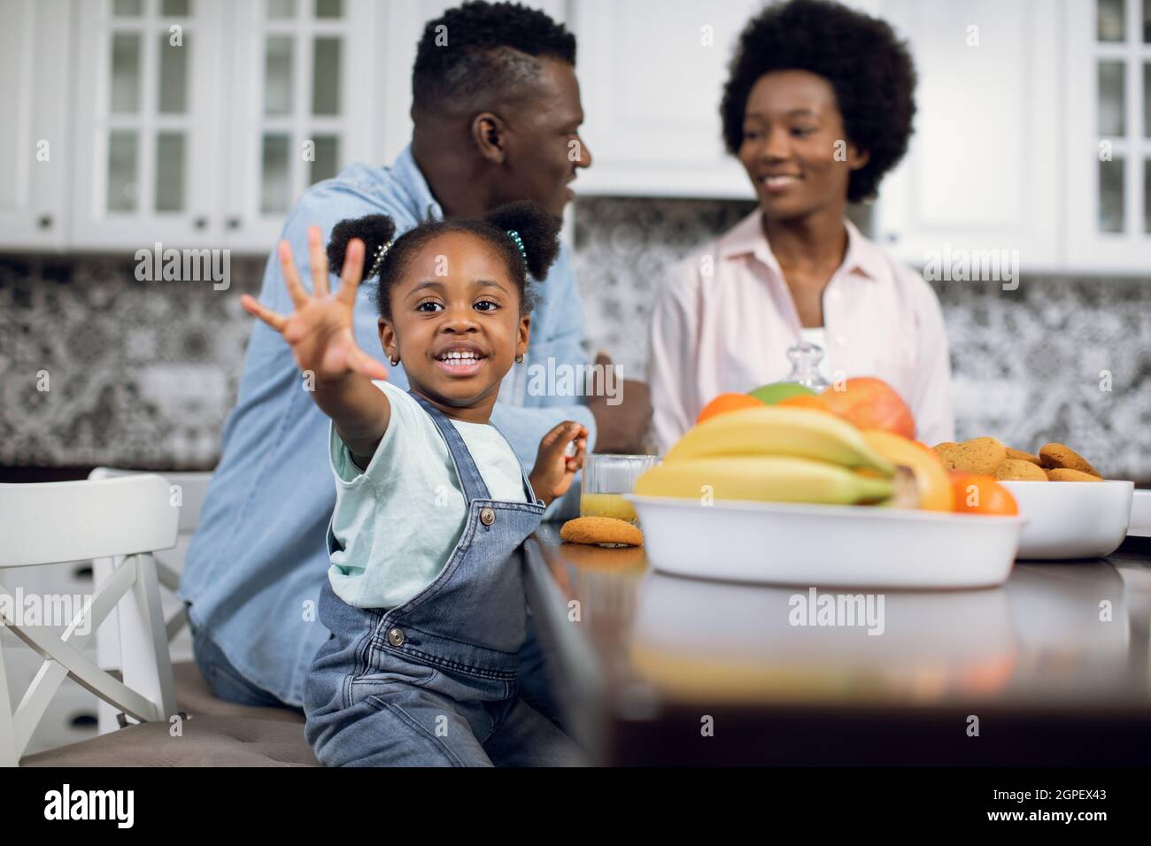 Pretty smiling girl waving hand on camera while sitting at modern kitchen with her parents. African wife and husband talking on blur background. Happy family spending time together at home. Stock Photo