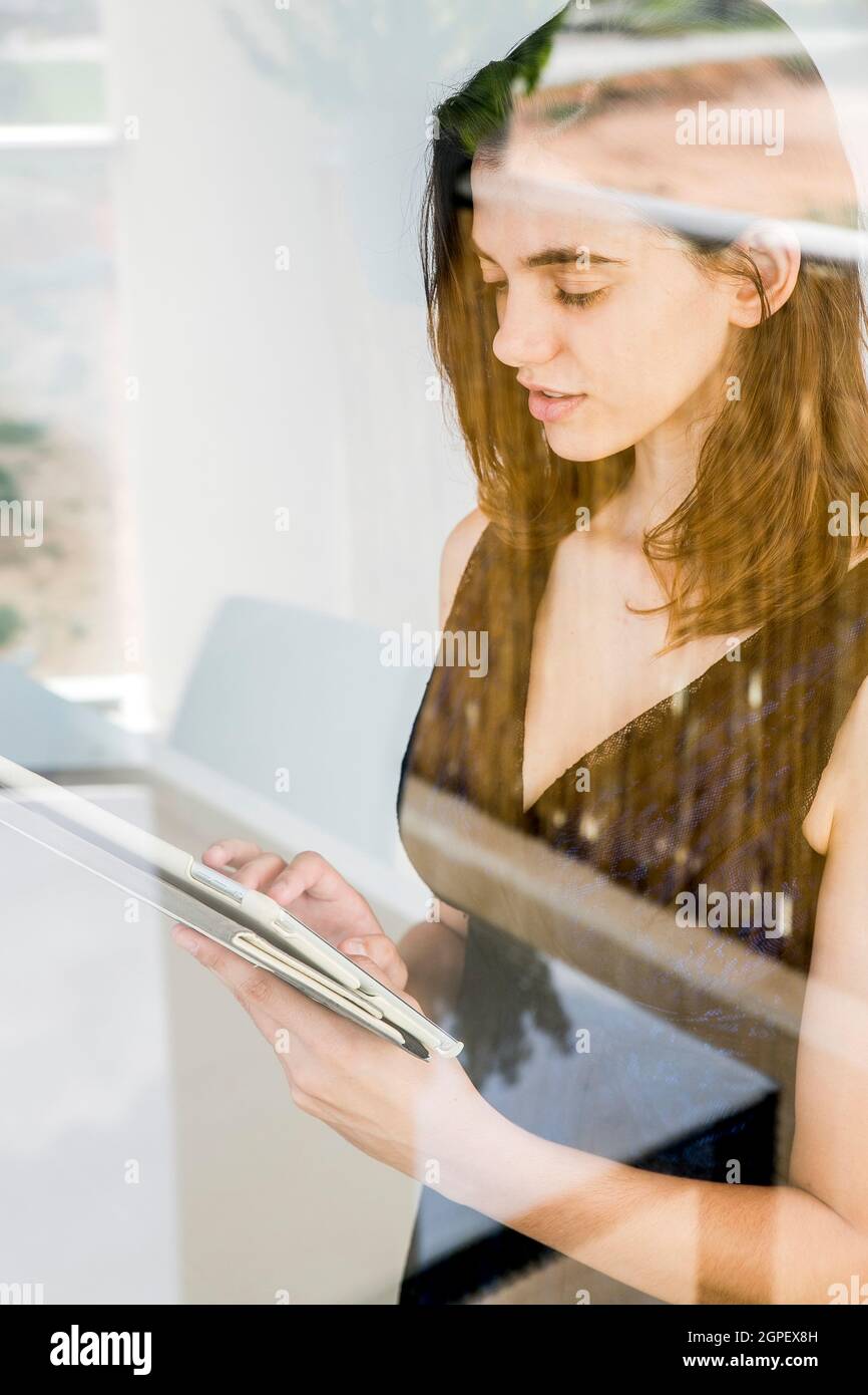 Young woman in formal dress works on her tablet at home as seen through the window Stock Photo