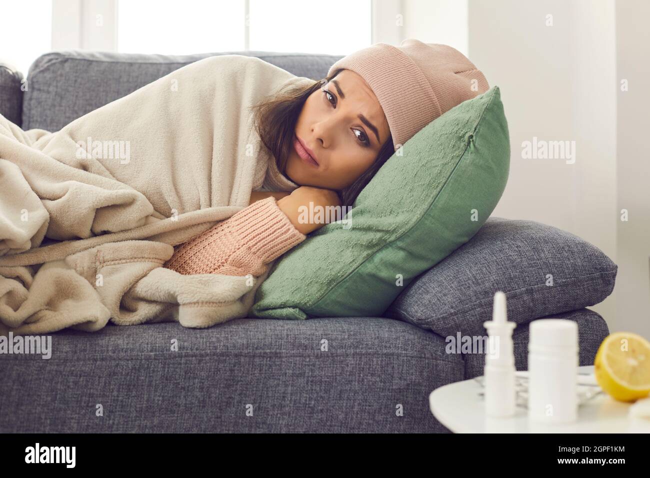 Young unhappy woman in warm clothes and hat lying in bed under blanket and feeling ill Stock Photo