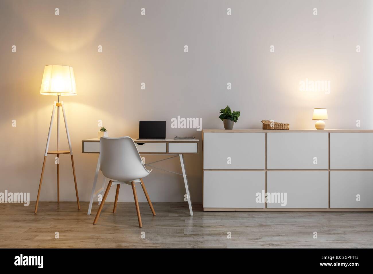 Cozy modern workplace in evening with table, computer, armchair and glowing lamp, on gray wall background Stock Photo