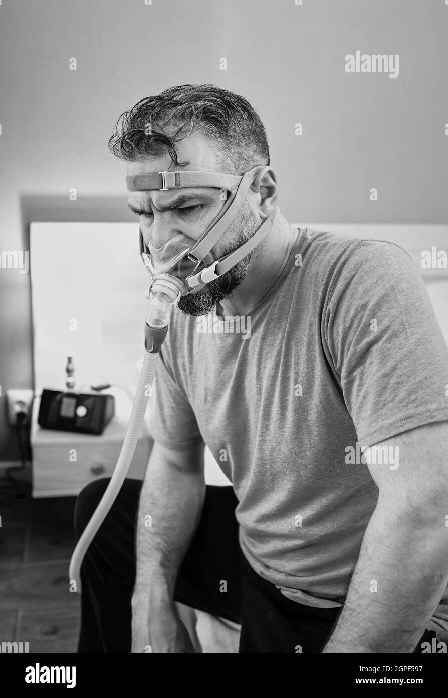 Unhappy shocked man with chronic breathing issues surprised by using  CPAP machine sitting on the bed in bedroom. Stock Photo