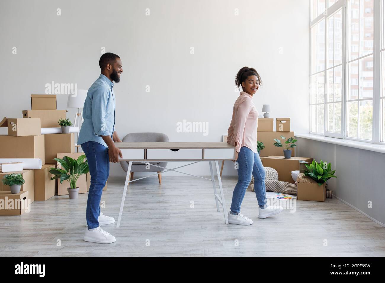 Cheerful young african american man and woman carry table in house with cardboard boxes, working together Stock Photo