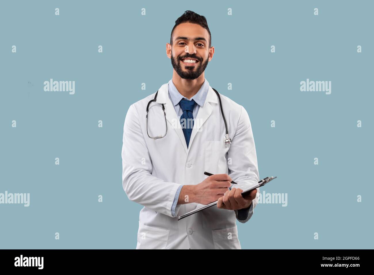 Happy Middle-Eastern Doctor Man Taking Notes Standing Over Blue Background Stock Photo