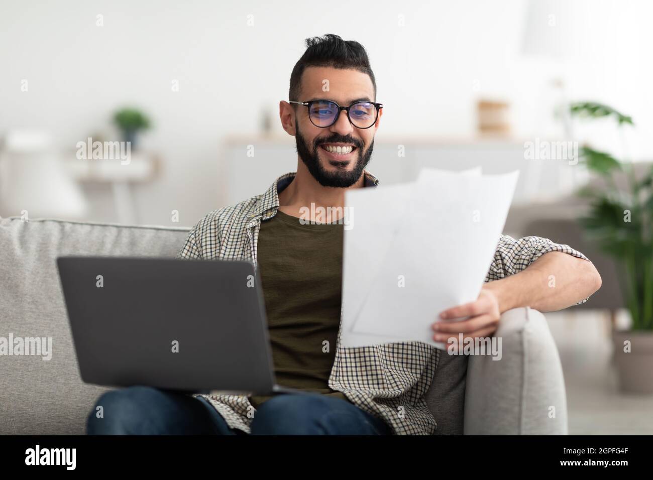 Young Arab guy making video call, reading documents, using laptop, working from home office Stock Photo