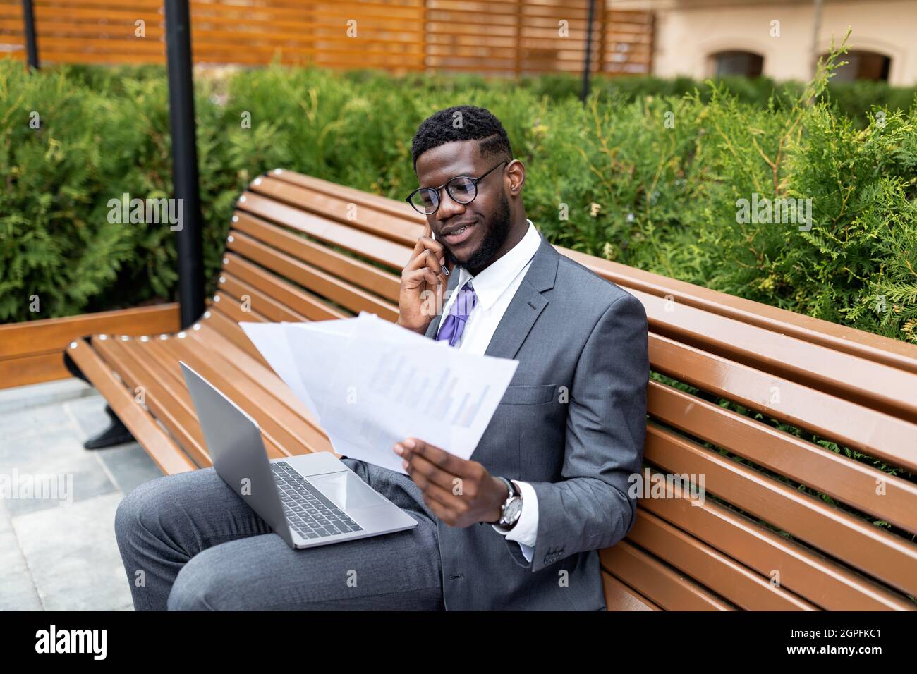 Busy lifestyle. Young african american businessman talking on cellphone, looking at documents, sitting on bench outdoors Stock Photo