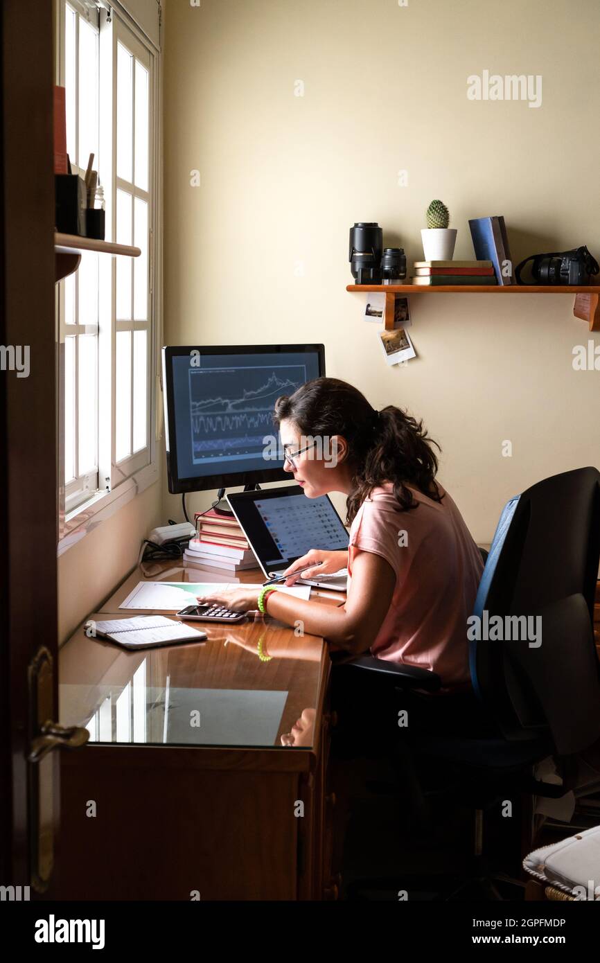 Brunette in glasses sitting at table with computer and investing in stock market during work remotely from home office Stock Photo
