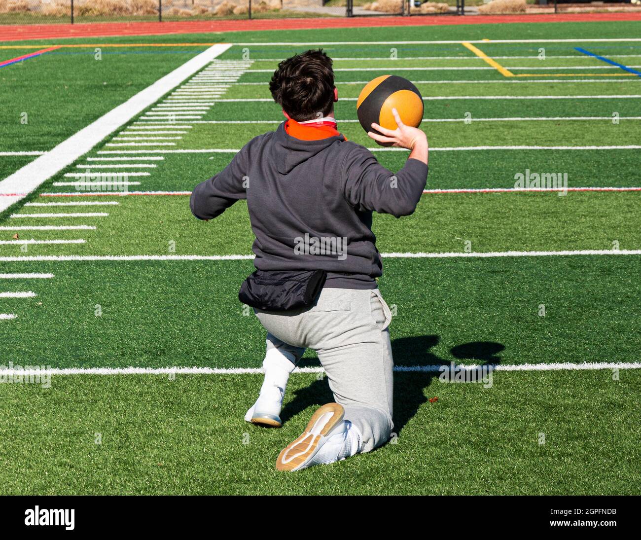 Rear view of a shot put track and field athlete on his knees training by throwing a medicine ball on a green turf field. Stock Photo