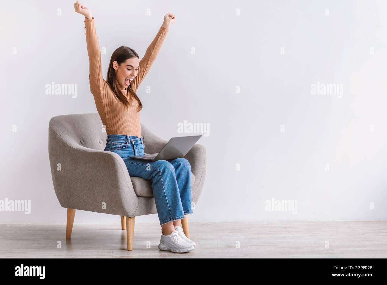 Excited young Caucasian lady sitting in armchair with laptop, lifting hands up, celebrating success against white wall Stock Photo
