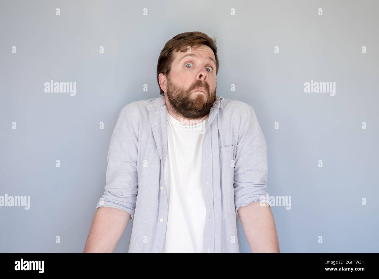 Bewilderment bearded man in a shirt shrugs in surprise and looks at the camera with a strange and funny expression on face.  Stock Photo