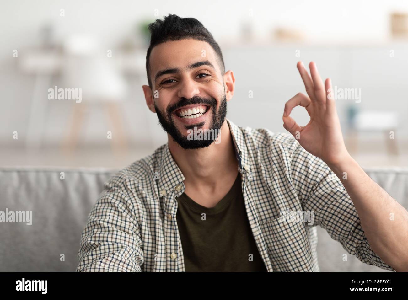 Happy young Arab guy showing okay gesture and smiling at camera, sitting on sofa at home Stock Photo