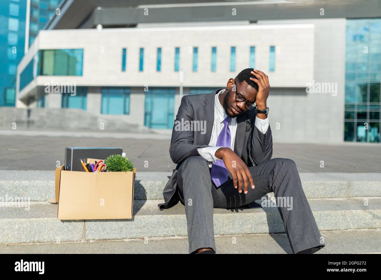Unemployment concept. Disappointed black businessman sitting with box of personal stuff on stairs against office center Stock Photo